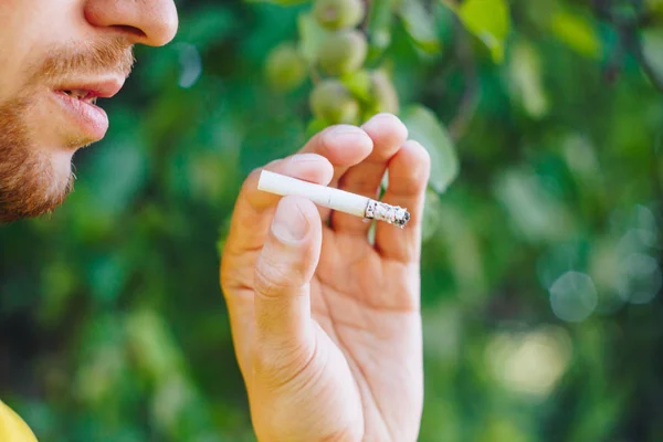 smoldering cigarette in the hand of a man with a beard on nature against the background of green trees. Nicotine tobacco smoke. Unhealthy Lifestyle. Close-up, ash. Smoking break