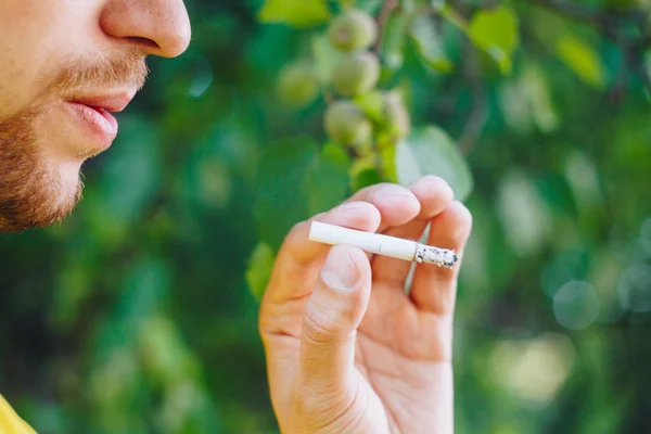 Cigarrillo humeante en la mano de un hombre con barba en la naturaleza sobre el fondo de árboles verdes. Humo de tabaco de nicotina. Estilo de vida insalubre. Primer plano, ceniza. Vacaciones para fumadores — Foto de Stock