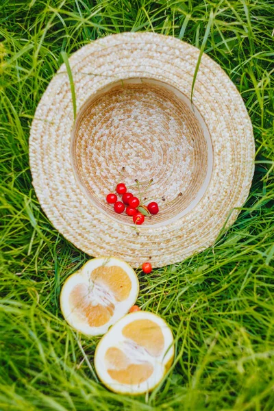 Cherry and cuted citrus fruits with straw hat lying on the grass outdoors. Picnic on nature in the park close up healthy food, diet, personal care. sunny day. — Stock Photo, Image