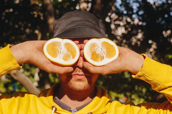 a man in a yellow hoodie and blue panama is holding two halves of a yellow citrus cut in half before his eyes. healthy eating, proper nutrition, organic food, close-up