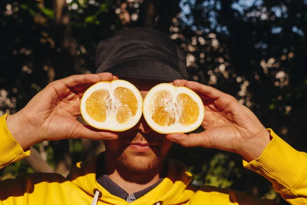 a man in a yellow hoodie and blue panama is holding two halves of a yellow citrus cut in half before his eyes. healthy eating, proper nutrition, organic food, close-up