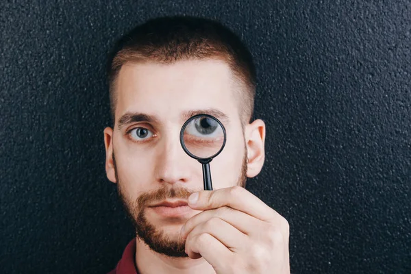 a young man with a beard looks through a magnifying glass. Portrait of a guy with a big eye on a black background. investigation, survey