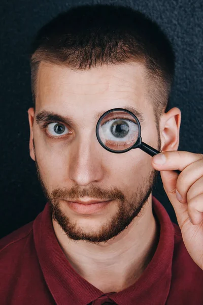 Un joven con barba mira a través de una lupa. Retrato de un tipo con un gran ojo en un fondo negro. investigación, encuesta — Foto de Stock