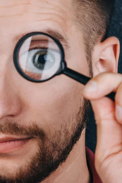 a young man with a beard looks through a magnifying glass. Portrait of a guy with a big eye on a black background. investigation, survey