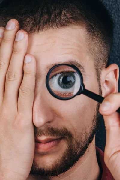 a young man with a beard looks through a magnifying glass. Portrait of a guy with a big eye on a black background. investigation, survey