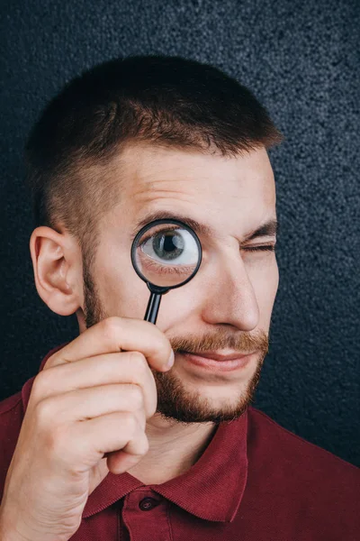 A young man with a beard looks through a magnifying glass. Portrait of a guy with a big eye on a black background. investigation, survey — Stock Photo, Image