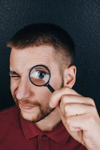 Un joven con barba mira a través de una lupa. Retrato de un tipo con un gran ojo en un fondo negro. investigación, encuesta —  Fotos de Stock