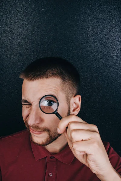 a young man with a beard looks through a magnifying glass. Portrait of a guy with a big eye on a black background. investigation, survey