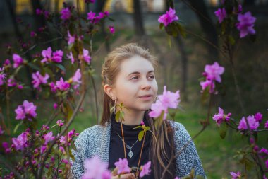 bulanık bir arka plan üzerinde bahar parkı pembe ve mor çiçeklerbir çalı içinde Avrupalı kadının açık hava portresi bir gülümseme ile uzak görünüyor