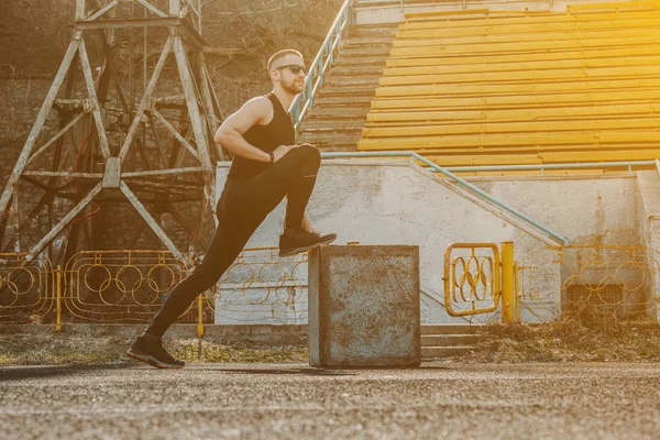 A slender young man in black clothes and sunglasses is exercising outdoors. fitness athlete on the sports field. training with projectiles. warm up stretching legs. body preparation for the summer