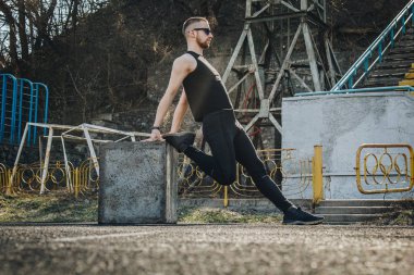 Siyah giysiler ve güneş gözlüğü ile ince bir genç adam açık havada egzersiz olduğunu. spor alanında fitness sporcusu. mermi ile eğitim. bacaklar germe kadar ısınmak. yaz için vücut hazırlığı