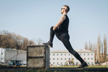Siyah giysiler ve güneş gözlüğü ile ince bir genç adam açık havada egzersiz olduğunu. spor alanında fitness sporcusu. mermi ile eğitim. bacaklar germe kadar ısınmak. yaz için vücut hazırlığı