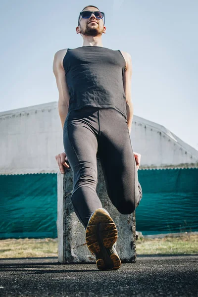 A slender young man in black clothes and sunglasses is exercising outdoors. fitness athlete on the sports field. training. vertical. warm up stretching legs. body preparation for the summer. vertical