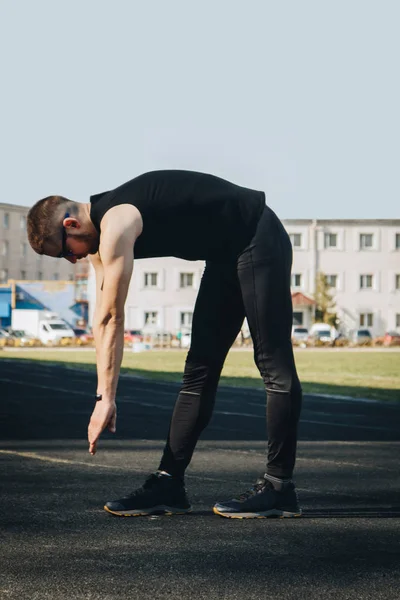 A slender young man in black clothes and sunglasses is exercising outdoors. fitness athlete on the sports field. training. vertical. warm up stretching legs. body preparation for the summer. vertical