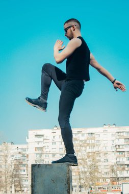 Siyah giysiler ve güneş gözlüğü ile ince bir genç adam açık havada egzersiz olduğunu. sahada fitness sporcusu. mermi ile eğitim. bacak eğitimi ısınmak. yaz için vücut hazırlığı. Dikey.