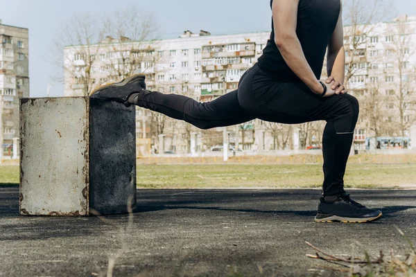 Smukły młody mężczyzna w czarnych ubraniach i okularów słonecznych wykonuje ćwiczenia na zewnątrz. sportowca fitness na boisku sportowym. Trening z pociskami. rozgrzać nogi rozciągające. przygotowanie ciała na lato — Zdjęcie stockowe