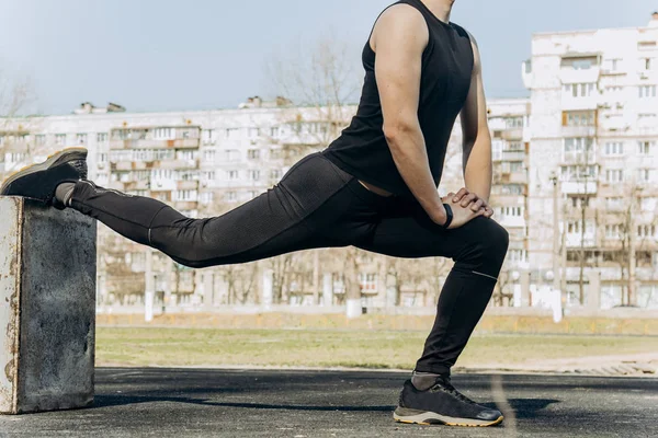 Smukły młody mężczyzna w czarnych ubraniach i okularów słonecznych wykonuje ćwiczenia na zewnątrz. sportowca fitness na boisku sportowym. Trening z pociskami. rozgrzać nogi rozciągające. przygotowanie ciała na lato — Zdjęcie stockowe