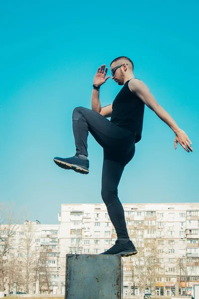 Siyah giysiler ve güneş gözlüğü ile ince bir genç adam açık havada egzersiz olduğunu. sahada fitness sporcusu. mermi ile eğitim. bacak eğitimi ısınmak. yaz için vücut hazırlığı. Dikey