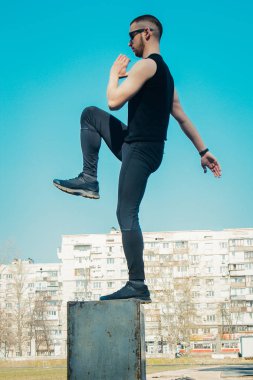 Siyah giysiler ve güneş gözlüğü ile ince bir genç adam açık havada egzersiz olduğunu. sahada fitness sporcusu. mermi ile eğitim. bacak eğitimi ısınmak. yaz için vücut hazırlığı. Dikey