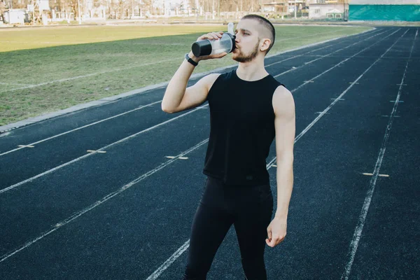 muscular slender man in training at the stadium drinks water from a sports shaker. recreation sports. athletic exercising