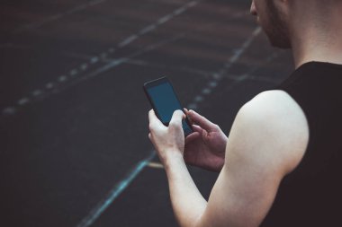 Siyah elbiseli genç bir adam telefona bakıyor. spor alanında fitness sporcusu. gadget'lar ile eğitim. yaz için vücut hazırlık ısınmak. izleme çalıştırma