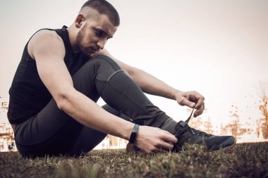 Siyah elbiseli genç bir adam, bağcıkları spor ayakkabılarına bağlıyor. çim üzerinde spor alanında oturan fitness sporcusu. eğitim için vücut hazırlık ısınma