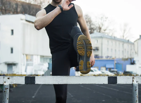 A man in black clothes is exercising outdoors with a barrier. fitness athlete on the sports field. training with hurdle. warm up stretching legs. body preparation for the summer