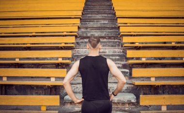 Siyah spor giyimli ince bir genç adam merdiven stadyum standları önünde duruyor. eylem için hazır, zafere adım, meydan okuma