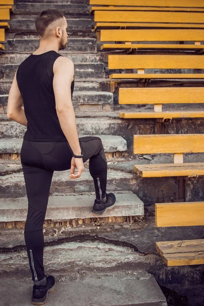 A slender young man in black sportswear stands in front of the stairs stadium stands. ready for action, step to victory, challenge. vertical