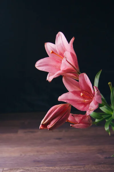 Strauß rosafarbener Lilienblüten in den Lichtstrahlen auf schwarzem Hintergrund auf einem rustikalen Holztisch. frische Knospen einer blühenden Pflanze aus nächster Nähe, Kopierraum. Studioaufnahmen. die Handlung der Urlaubskarte — Stockfoto