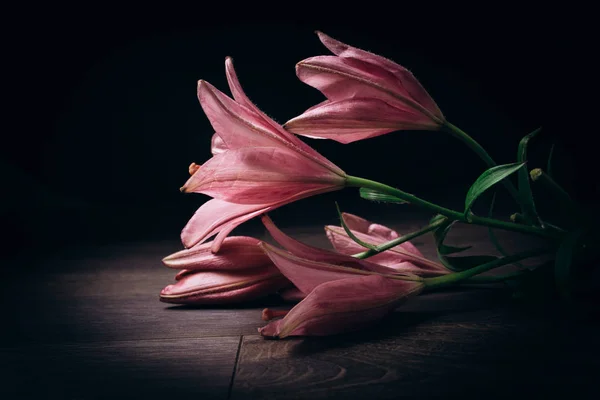 Strauß rosafarbener Lilienblüten in den Lichtstrahlen auf schwarzem Hintergrund auf einem rustikalen Holztisch. frische Knospen einer blühenden Pflanze aus nächster Nähe, Kopierraum. Studioaufnahmen. die Handlung der Urlaubskarte — Stockfoto