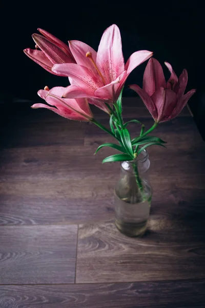 Ramo de flores de lirio rosa en los rayos de luz sobre un fondo negro sobre una mesa rústica de madera en una botella de vidrio. los cogollos frescos de la planta que florece se acercan en el florero, copian el espacio. plano de estudio — Foto de Stock