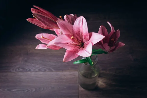 Strauß rosa Lilienblüten in den Lichtstrahlen auf schwarzem Hintergrund auf einem rustikalen Holztisch in einer Glasflasche. frische Knospen einer blühenden Pflanze in Großaufnahme in einer Vase, Kopierraum. Studioaufnahme — Stockfoto