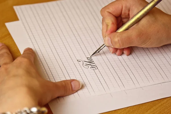 Vrouwelijke hand schrijft met een inkt pen op een wit papier blad met strepen. Briefpapier op houten bureau close-up top View. spelling lessen en caligraphy oefeningen — Stockfoto