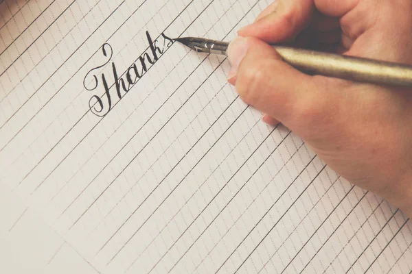 Female hand writes with the inky pen the word thanks on a white paper sheet with stripes. stationery on desk close up top view. spelling lessons and caligraphy exercises. Template, layout, background — Stock Photo, Image