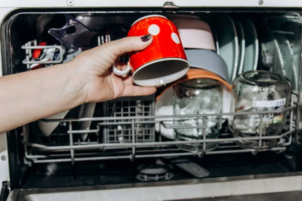 Female hand lays the dishes in an open dishwasher close-up clogged with clean, washed dishes. dry cutlery closeup. spoons forks. mugs, plates. household appliances in the kitchen