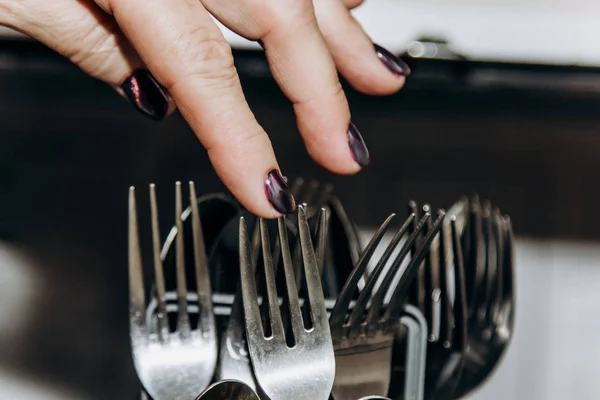Female hand lays the dishes in an open dishwasher close-up clogged with clean, washed dishes. dry cutlery closeup. spoons forks. mugs, plates. household appliances in the kitchen — Stock Photo, Image