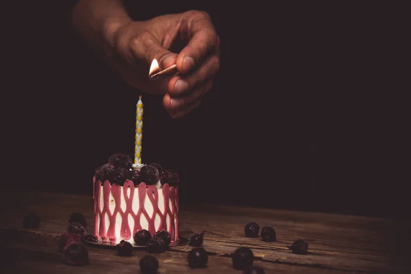 Mano enciende una vela un pastel festivo con los frutos de la cereza en una mesa de madera rústica sobre un fondo oscuro. cerrar espacio de copia. patrón vintage en tartaleta de postre. vacaciones de cumpleaños —  Fotos de Stock