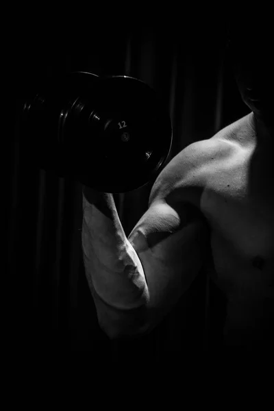 Atleta entrena bíceps manos con mancuernas en el centro de los entrenamientos sobre un fondo negro. herramientas de entrenamiento en el primer plano del gimnasio —  Fotos de Stock