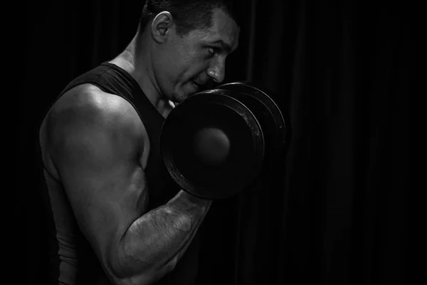 Atleta entrena bíceps manos con mancuernas en el centro de los entrenamientos sobre un fondo negro. herramientas de entrenamiento en el primer plano del gimnasio —  Fotos de Stock