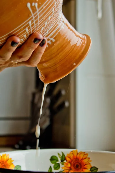 Жіноча рука наливає молоко з керамічної плити в металеву миску. пофарбований у білу емаль, коричневий глиняний глечик крупним планом. вертикальний — стокове фото