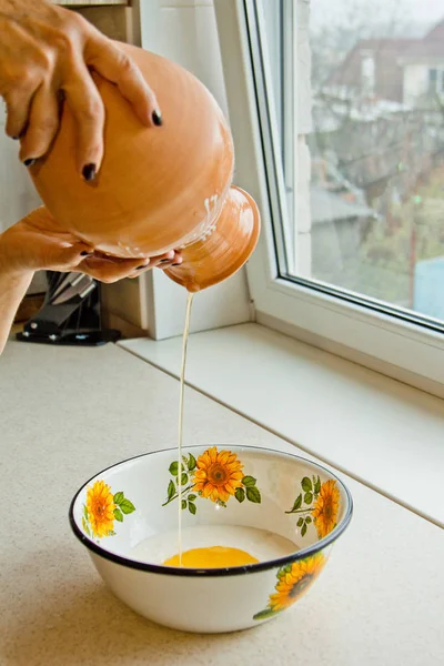 Mão feminina derramando óleo de cerâmica ewer em uma tigela de metal. pintado em esmalte branco, um jarro de barro marrom close-up vertical. massa de farinha de cozinha — Fotografia de Stock