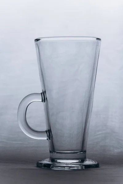 Taza de vidrio transparente vacía sobre un fondo gris plateado de cerca. taza alta con mango y pierna. espacio de copia, textura, fondo. vertical — Foto de Stock