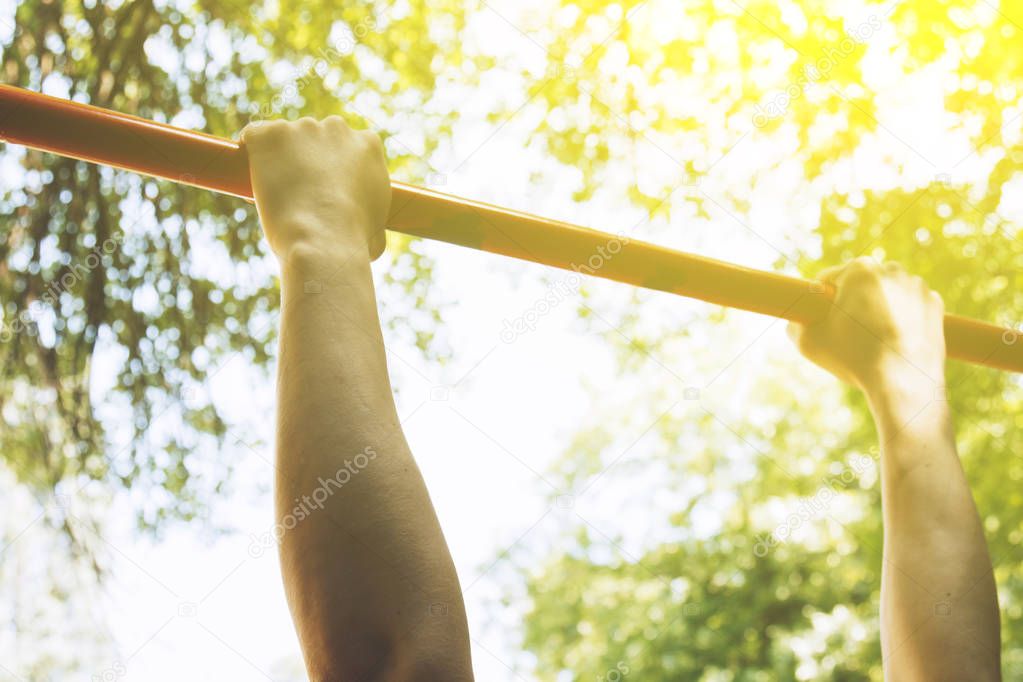 the hands of a sports athlete hold on to a metal crossbar for pull-ups on the training ground against a clear sky and green foliage. sports training outdoors. workout first-person view