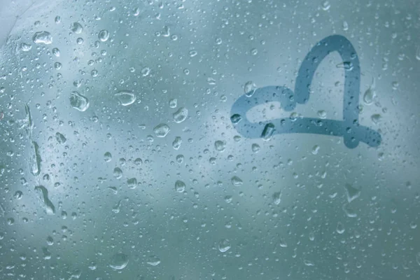 手指在半透明的汗玻璃上画出心形的爱的象征。春雨雨滴在窗户特写上。模糊的背景复制空间 — 图库照片