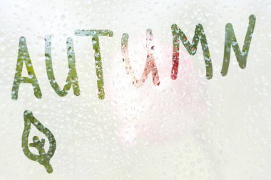 Yarı saydam, sisli bir cam üzerinde sonbahar kelimesinin şeritlerinin şeklinin bir parmak çizimi. pencere yakın çekim bahar yağmur damlaları. bulanık arka plan kopyalama alanı