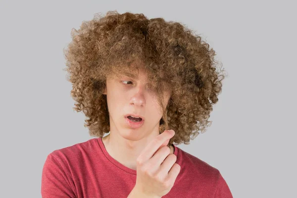Portrait of a young curly european man frightened looks at his long hair. holds a curl of hair with his fingers. very lush male guy hair. curling hair for men. isolated on gray background — Stock Photo, Image