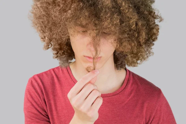 Retrato de um jovem europeu encaracolado olhando preocupadamente para seu cabelo longo. segura um fio de cabelo com os dedos. cabelo masculino muito exuberante cara. cabelo ondulação para os homens. isolado em fundo cinza — Fotografia de Stock