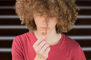 Genç kıvırcık Avrupalı bir adamın pencereye karşı uzun saçlarına endişeyle baktığı bir portresi. parmaklarıyla bir saç kıvrımı tutar. çok gür erkek adam saç. erkekler için saç kıvırma