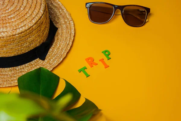 Αξεσουάρ παραλίας: ψάθινο καπέλο, γυαλιά ηλίου, το ταξίδι επιγραφή και ένα πλατύ φύλλο τέρας ένα φυτό για σκιά σε ένα φωτεινό κίτρινο φόντο σε μια ηλιόλουστη μέρα, επάνω όψη, αντίγραφο χώρου — Φωτογραφία Αρχείου